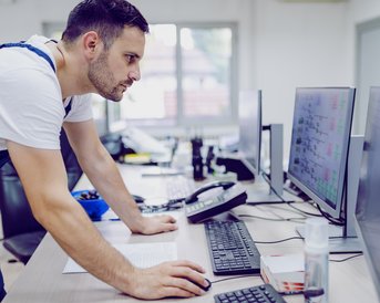 Ein Mann in Arbeitskleidung steht an einem Schreibtisch und schaut konzentriert auf mehrere Computerbildschirme. Der Raum ist hell und modern eingerichtet.