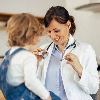 Eine Ärztin in einem weißen Kittel hält ein Stethoskop und lächelt, während sie mit einem kleinen Kind spricht, das auf ihrem Schoß sitzt.