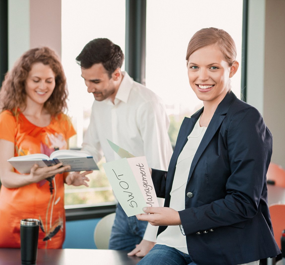 Drei Personen stehen in einem modernen Büro. Eine Frau hält ein Blatt mit der Aufschrift "SWOT Management", während die anderen beiden in ein Buch schauen. Helle Fenster bieten einen Blick auf die Außenansicht.