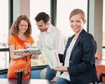 Drei Personen stehen in einem modernen Büro. Eine Frau hält ein Blatt mit der Aufschrift "SWOT Management", während die anderen beiden in ein Buch schauen. Helle Fenster bieten einen Blick auf die Außenansicht.