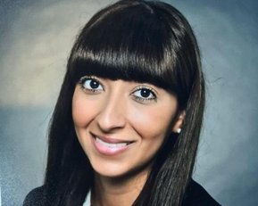 Eine Frau mit langen, glatten, braunen Haaren und einem geraden Pony lächelt in die Kamera. Sie trägt ein schwarzes Blazer-Jackett und ein weißes Hemd. Der Hintergrund ist unscharf und neutral.
