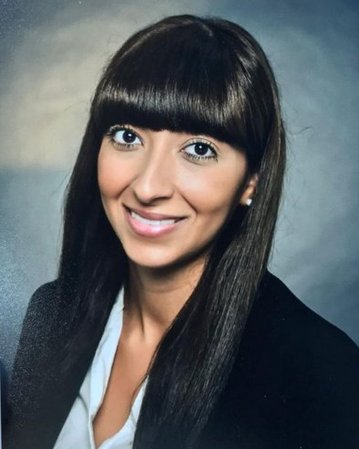 Eine Frau mit langen, glatten, braunen Haaren und einem geraden Pony lächelt in die Kamera. Sie trägt ein schwarzes Blazer-Jackett und ein weißes Hemd. Der Hintergrund ist unscharf und neutral.