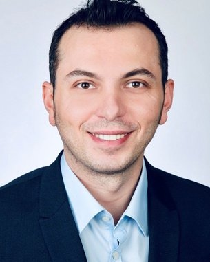 Ein Mann mit dunklem Haar und einem freundlichen Lächeln trägt einen Anzug und ein hellblaues Hemd. Der Hintergrund ist neutral.