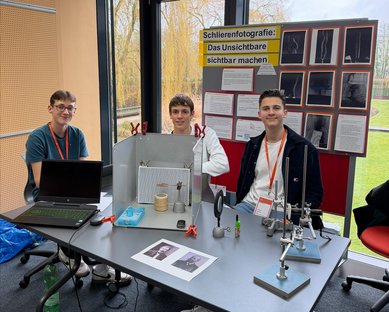 Drei Schüler sitzen an einem Tisch mit einem Laptop und Experimenten zur Schlierenfotografie. Im Hintergrund sind Poster mit Informationen und Bildern.