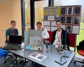Three students are sitting at a table with a laptop and experiments on schlieren photography. In the background, there are posters with information and images.