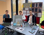 Drei Schüler sitzen an einem Tisch mit einem Laptop und Experimenten zur Schlierenfotografie. Im Hintergrund sind Poster mit Informationen und Bildern.