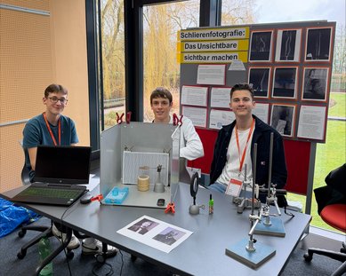 Three students are sitting at a table with a laptop and experiments on schlieren photography. In the background, there are posters with information and images.