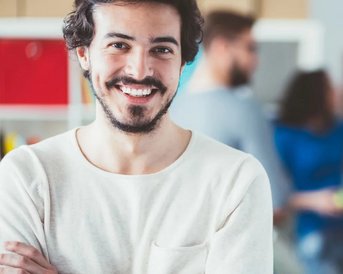 A young man with a broad smile stands in the foreground. He is wearing a light sweater and has dark, curly hair. In the background, there are blurred figures interacting with each other.