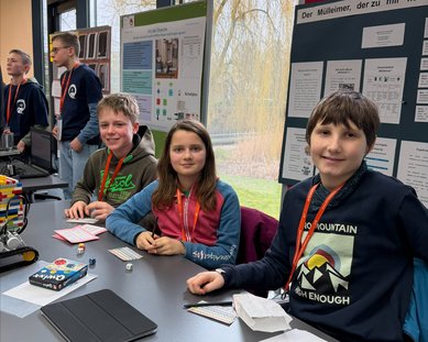 Four children are sitting at a table, surrounded by project posters. They are working on a robot and wearing name tags around their necks.