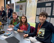 Four children are sitting at a table, surrounded by project posters. They are working on a robot and wearing name tags around their necks.
