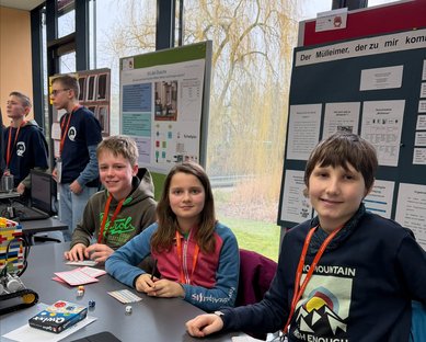 Vier Kinder sitzen an einem Tisch, umgeben von Projektplakaten. Sie arbeiten an einem Roboter und haben Namensschilder um den Hals.