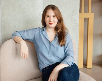 Eine Frau mit langen, braunen Haaren sitzt auf einem beigen Sessel und trägt ein hellblaues Hemd. Neben ihr steht ein kleiner Tisch mit einem Glas Milch.