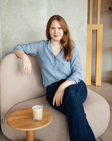 Eine Frau mit langen, braunen Haaren sitzt auf einem beigen Sessel und trägt ein hellblaues Hemd. Neben ihr steht ein kleiner Tisch mit einem Glas Milch.