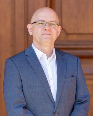 Mann in Anzug mit Brille steht vor einer Holzoberfläche. Er hat eine glatte Kopfhaut und trägt ein weißes Hemd unter dem Anzug.