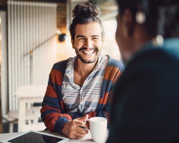 Ein lächelnder Mann mit lockigem Haar sitzt an einem Tisch, hält eine Tasse und spricht mit einer Person außerhalb des Rahmens. Auf dem Tisch liegt ein Tablet. Der Hintergrund zeigt eine moderne, helle Umgebung.