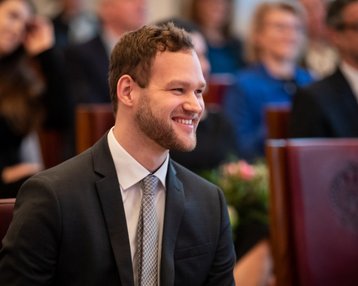 Ein junger Mann in einem Anzug sitzt lächelnd in einer Versammlung, umgeben von anderen Anwesenden in einem festlichen Raum.