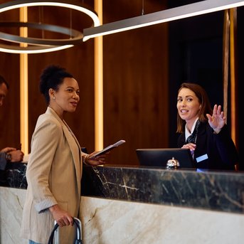 Eine Frau mit einem Regenschirm spricht an der Rezeption mit einer Mitarbeiterin, während ein Mann im Hintergrund an einem Tisch arbeitet.