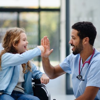 Ein Arzt gibt einem Mädchen im Rollstuhl ein High-Five. Beide lächeln, während eine weitere Person im Hintergrund steht.