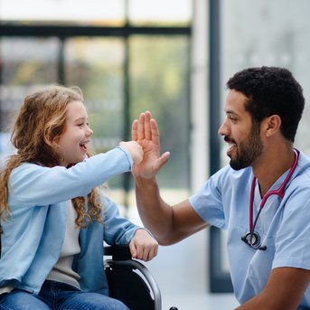 Ein Arzt gibt einem Mädchen im Rollstuhl ein High-Five. Beide lächeln, während eine weitere Person im Hintergrund steht.