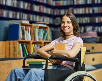 Eine junge Frau im Rollstuhl sitzt lächelnd in einer Bibliothek und hält Bücher in den Händen. Im Hintergrund sind Regale mit Büchern sichtbar.