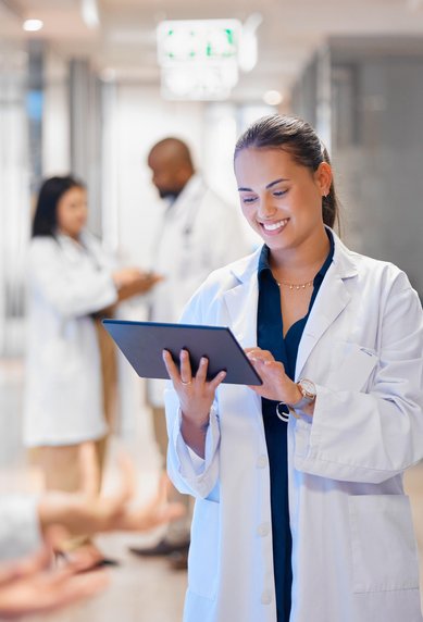 Eine Ärztin in einem weißen Kittel hält ein Tablet und lächelt, während sie in einem modernen Krankenhausflur steht. Im Hintergrund sind weitere Personen zu sehen.