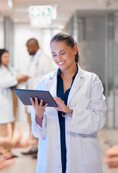 Eine Ärztin in einem weißen Kittel hält ein Tablet und lächelt, während sie in einem modernen Krankenhausflur steht. Im Hintergrund sind weitere Personen zu sehen.