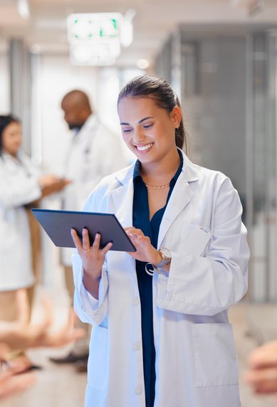 Eine Ärztin in einem weißen Kittel hält ein Tablet und lächelt, während sie in einem modernen Krankenhausflur steht. Im Hintergrund sind weitere Personen zu sehen.