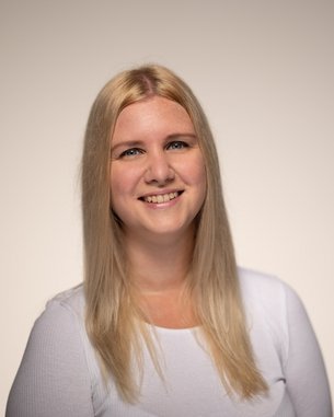 A smiling woman with long blonde hair is wearing a white top. The background is neutral and bright.