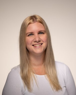 Eine lächelnde Frau mit langen, blonden Haaren trägt ein weißes Oberteil. Der Hintergrund ist neutral und hell.