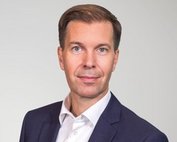 Ein Mann in einem Anzug mit einem weißen Hemd und einer weißen Tasche auf der linken Seite, vor einem neutralen Hintergrund.