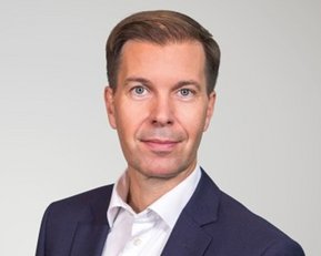 Ein Mann in einem Anzug mit einem weißen Hemd und einer weißen Tasche auf der linken Seite, vor einem neutralen Hintergrund.