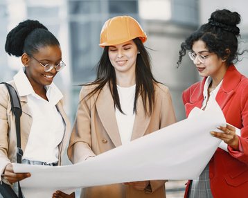 Kategorie Ingeniurwissenschaften SRH University Drei Frauen betrachten gemeinsam Baupläne, während sie in einem modernen urbanen Umfeld stehen. Eine Frau trägt einen orangefarbenen Helm, während die anderen Brillen tragen und lächeln.