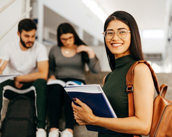 A smiling student with glasses is holding notebooks while sitting on a staircase. In the background, two other students are looking at books.