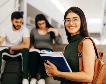 Eine lächelnde Studentin mit Brille hält Notizbücher in der Hand, während sie auf einer Treppe sitzt. Im Hintergrund sind zwei weitere Studenten, die in Bücher schauen.