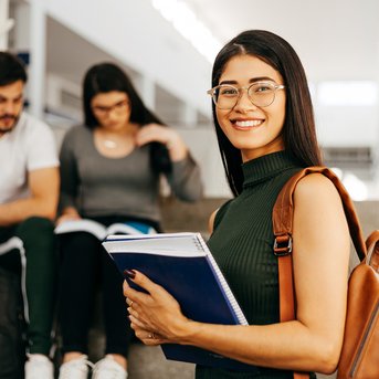 A smiling student with glasses is holding notebooks while sitting on a staircase. In the background, two other students are looking at books.