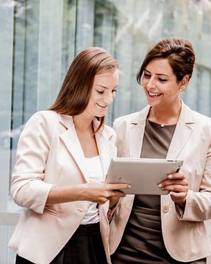 Zwei Frauen in formeller Kleidung stehen draußen und betrachten gemeinsam ein Tablet. Eine Frau zeigt auf den Bildschirm, während die andere aufmerksam zuschaut. Im Hintergrund sind moderne Gebäude und Bäume sichtbar.