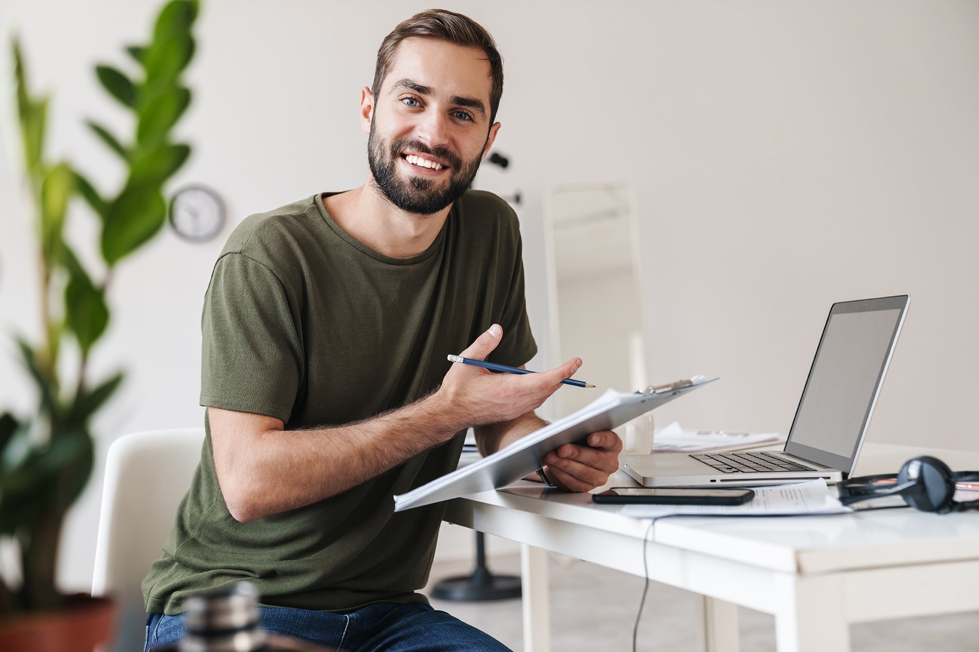 A young man is sitting at a table with a laptop and a notepad. He is holding a pen in his hand and smiling as he looks at the camera. In the background, there are plants and a clock visible.