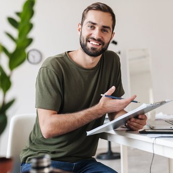 Ein junger Mann sitzt an einem Tisch mit einem Laptop und einem Notizblock. Er hält einen Stift in der Hand und lächelt, während er auf die Kamera schaut. Im Hintergrund sind Pflanzen und eine Uhr sichtbar.