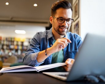 Ein Mann sitzt an einem Tisch in einem modernen Raum, arbeitet an einem Laptop und hält einen Stift in der Hand. Vor ihm liegt ein aufgeschlagenes Buch. Er trägt eine Brille und hat einen lässigen Kleidungsstil.