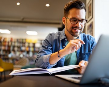 Ein Mann sitzt an einem Tisch in einem modernen Raum, arbeitet an einem Laptop und hält einen Stift in der Hand. Vor ihm liegt ein aufgeschlagenes Buch. Er trägt eine Brille und hat einen lässigen Kleidungsstil.