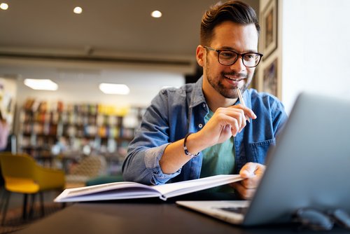 Ein Mann sitzt an einem Tisch in einem modernen Raum, arbeitet an einem Laptop und hält einen Stift in der Hand. Vor ihm liegt ein aufgeschlagenes Buch. Er trägt eine Brille und hat einen lässigen Kleidungsstil.