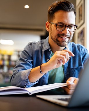 Ein Mann sitzt an einem Tisch in einem modernen Raum, arbeitet an einem Laptop und hält einen Stift in der Hand. Vor ihm liegt ein aufgeschlagenes Buch. Er trägt eine Brille und hat einen lässigen Kleidungsstil.