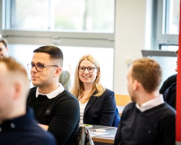Eine Gruppe von Menschen sitzt in einem Raum. Eine Frau mit Brille und blonden Haaren lächelt, während andere Teilnehmer in ihre Richtung schauen.