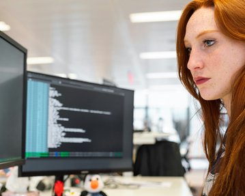 A woman with red hair is intently looking at two computer screens displaying programming code.