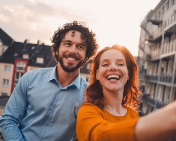 Ein Mann mit lockigem Haar und eine Frau mit langen roten Haaren lächeln, während sie ein Selfie auf einem Balkon machen. Im Hintergrund sind Wohngebäude und ein Sonnenuntergang sichtbar.