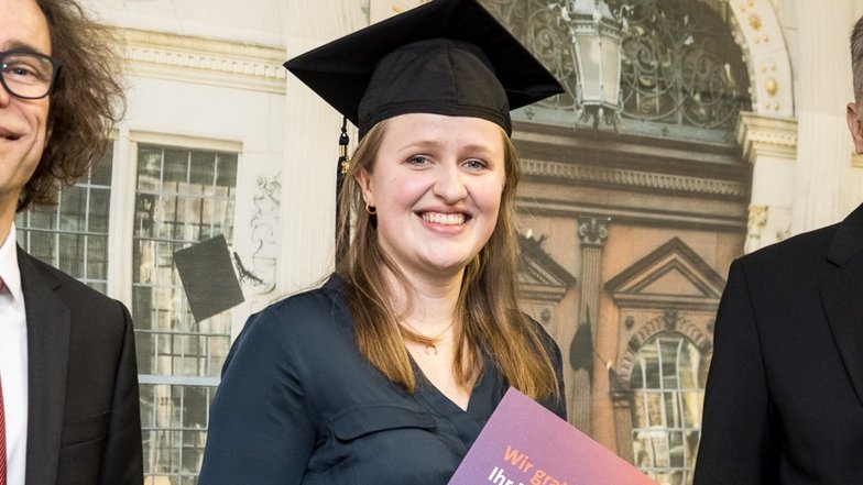 Eine graduierte Frau in einem schwarzen Hut hält ein Diplom in der Hand, umgeben von zwei Männern in formeller Kleidung.