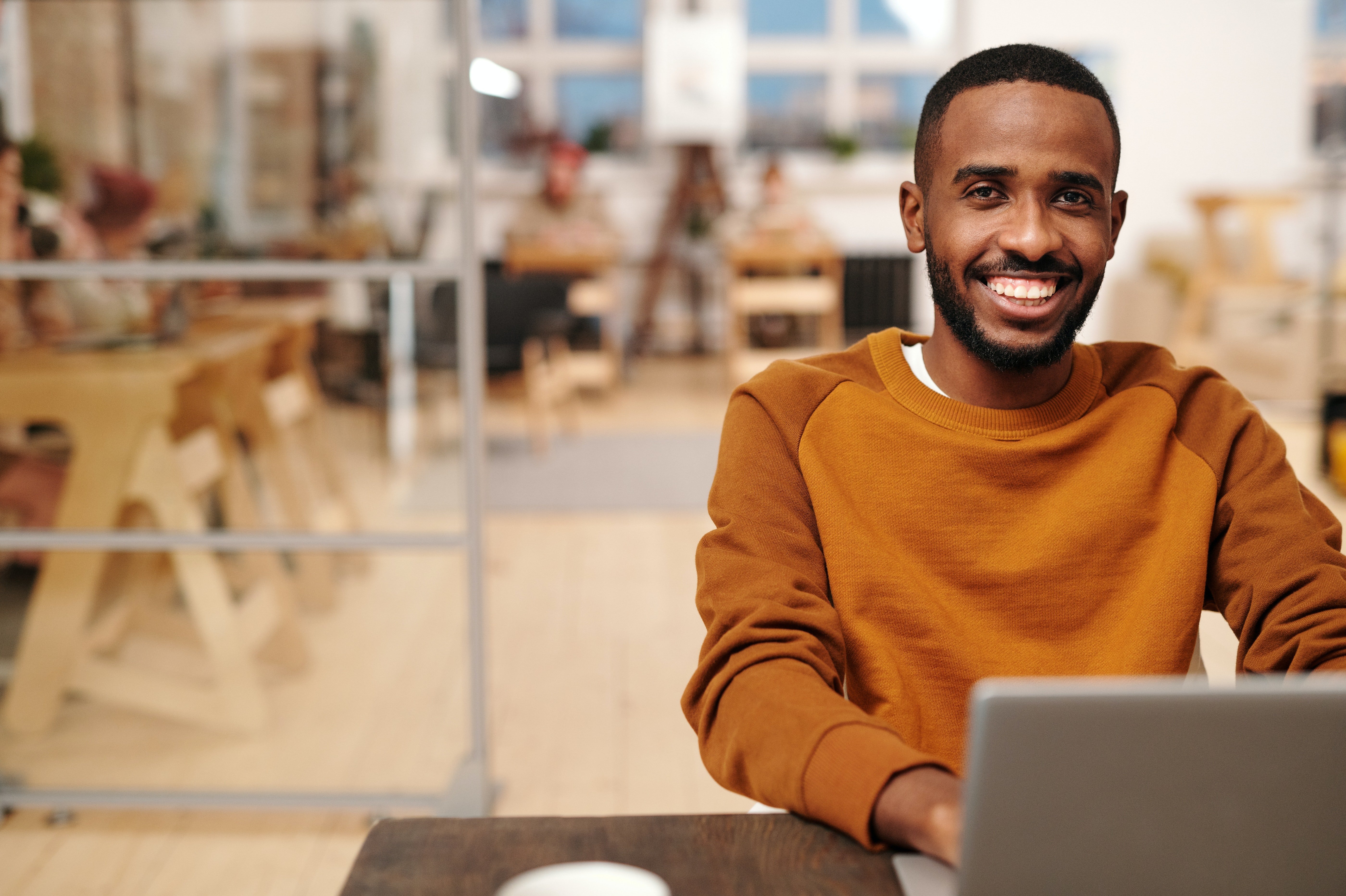 A young man with a broad smile sits at a table working on a laptop. He is wearing an orange sweater and is in a modern, bright café with wooden interiors.