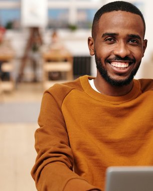 Ein junger Mann mit einem breiten Lächeln sitzt an einem Tisch und arbeitet an einem Laptop. Er trägt einen orangefarbenen Pullover und befindet sich in einem modernen, hellen Café mit Holzinterieur.