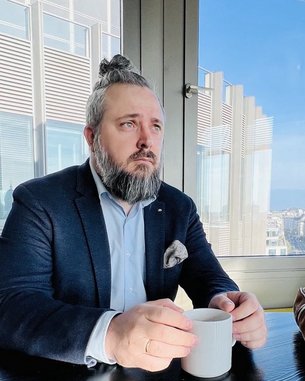 Ein Mann mit Bart und Zopf sitzt an einem Tisch und hält eine Tasse in der Hand, während er aus dem Fenster schaut.