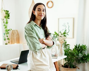 A woman with long, dark hair stands in a bright, modern office with plants and a laptop on the table.